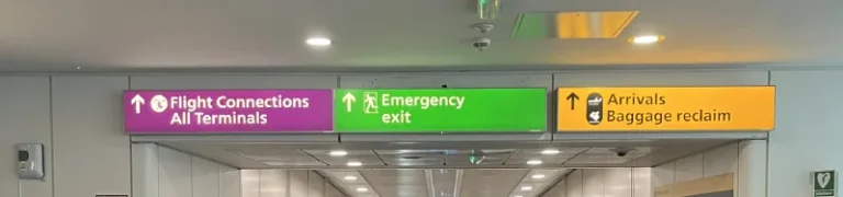 Overhead signs inside a Heathrow terminal: a purple Flight Connections sign for all terminals, a green emergency exit sign and an amber sign for arrivals and baggage reclaim