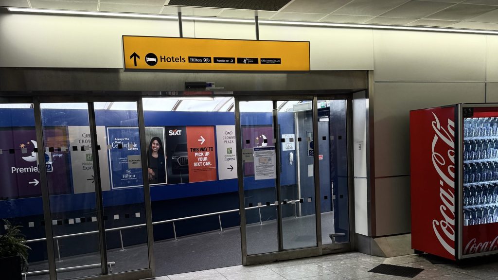Yellow Hotels sign at Heathrow Terminal 4 above the glass doors to the covered walkway, listing Hilton, Premier Inn, Holiday Inn Express and Crowne Plaza